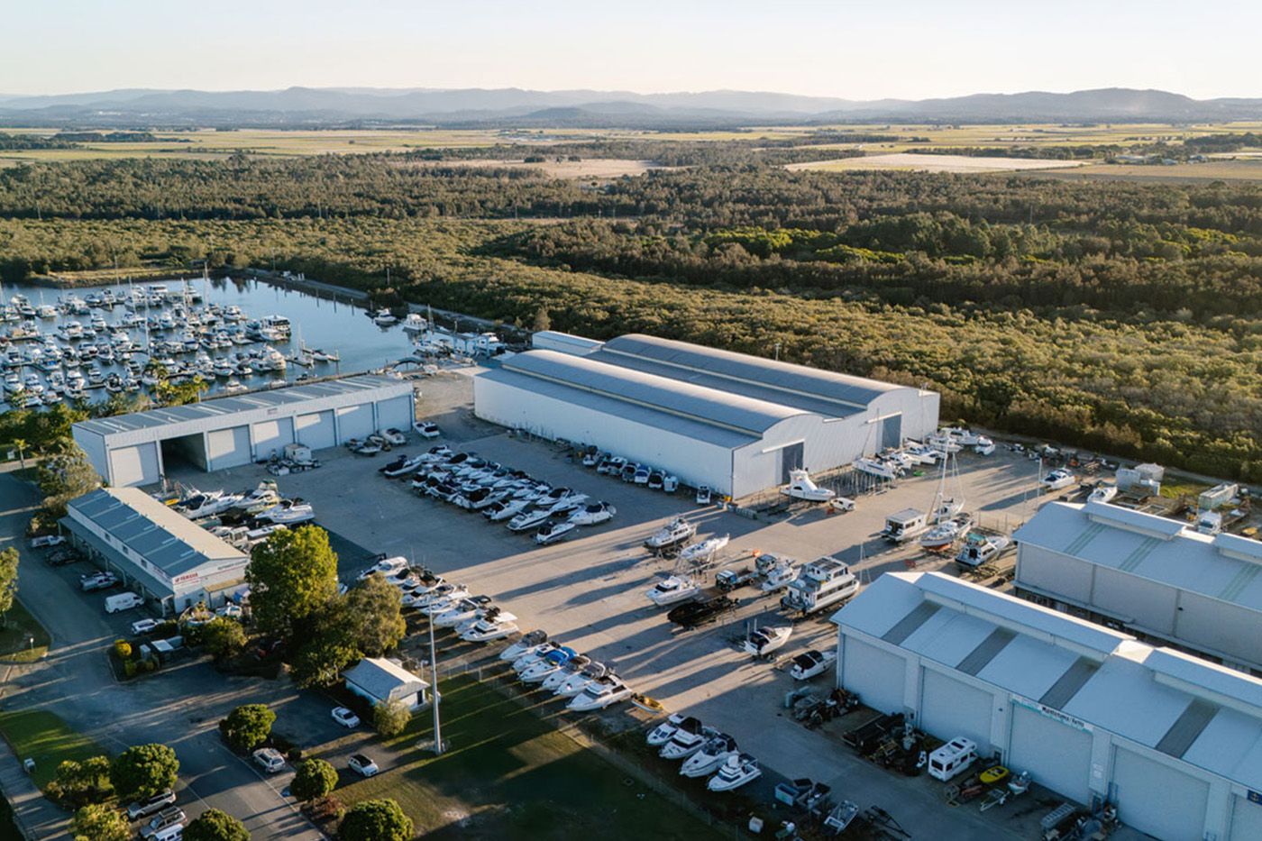 Aerial view of Pacific Point Marina & Boatyard: marina berths beside large boat sheds and a hardstand lined with boats, with bushland and the hinterland behind